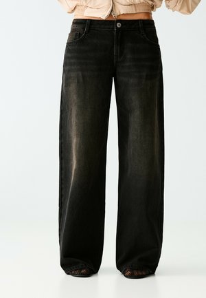 Jean noir à jambes larges porté par une personne avec une veste courte beige et des chaussures noires à bout ouvert avec des chaussettes en résille.