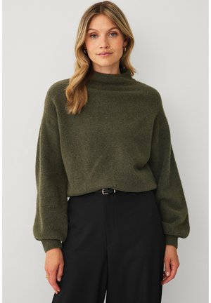 Kvinde med bølget blondt hår iført en mørkegrøn sweater med ballonærmer og høj hals, sorte bukser, stående mod en ensfarvet hvid baggrund.