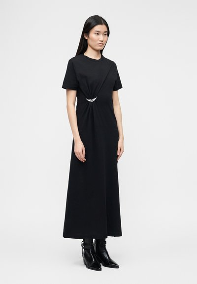 Γυναίκα που φοράει μαύρο μακρύ φόρεμα με κοντά μανίκια και λεπτομέρεια στη συγκέντρωση μπροστά, καθώς και μαύρα μπότες αστραγάλου με μυτερά δάχτυλα, στέκεται μπροστά σε απλό φόντο.