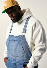 Mand iført sennepsgul kasket, hvid hoodie og blå denimoveraller, der kigger mod kameraet med hænderne i lommerne.