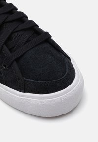 Black suede sneaker with textured upper, flat black laces, and white rubber sole. Features a rounded toe and contrast stitching details.
