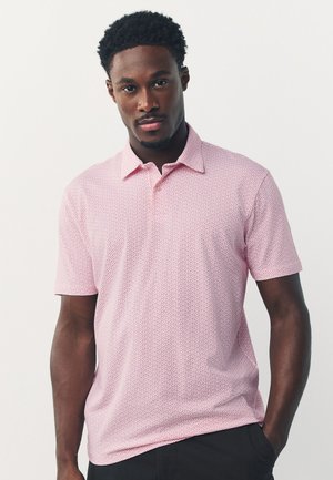 Homme aux cheveux courts et bouclés portant un polo rose clair à manches courtes et motifs, ainsi qu’un pantalon noir sur un fond uni.