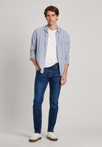 Camisa de botones a rayas azul claro y blanca, camiseta blanca, jeans azul oscuro y zapatillas blancas con suelas marrones.