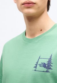 Κοντινό πλάνο ενός ατόμου που φορεί πράσινο μπλουζάκι με γραφικό σχέδιο μπλε σκηνής και δέντρου, και το λογότυπο "The North Face" στο στήθος.