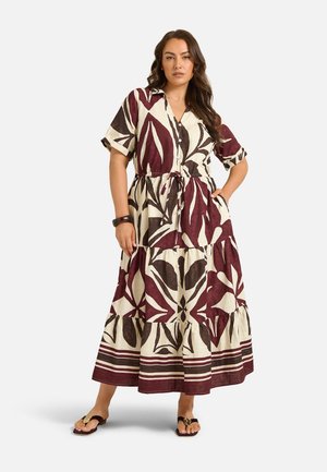 Femme portant une longue robe crème avec de grands motifs de feuilles rouge foncé et marron, manches courtes, boutons et taille à cordon, associée à des sandales.
