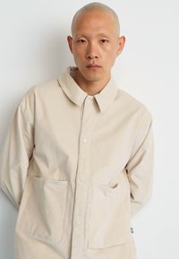 Camicia beige con bottoni e colletto, realizzata in tessuto leggero. Presenta due tasche frontali e un motivo sottile e strutturato.
