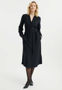 Vestido negro de manga larga con cinturón, escote en V y bolsillos. Tejido suave con corte recto, largo hasta la rodilla y aberturas laterales.