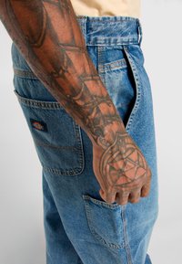 Blue denim cargo pants with multiple pockets and visible stitching. Hand with intricate tattoos rests against the side of the pants.