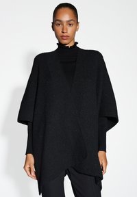 Zwarte gebreide poncho met korte mouwen, voorzien van een diepe V-hals en een asymmetrische zoom. De stof is getextureerd met subtiele glinsteraccenten.
