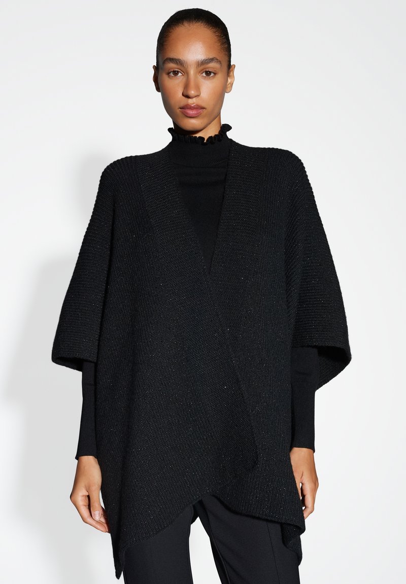 Zwarte gebreide poncho met korte mouwen, voorzien van een diepe V-hals en een asymmetrische zoom. De stof is getextureerd met subtiele glinsteraccenten.