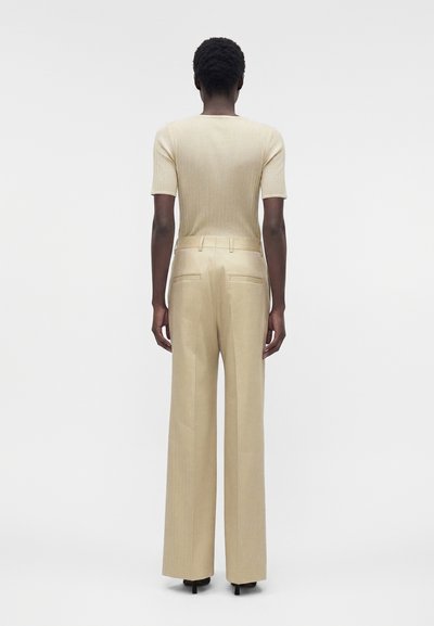 Άτομο που στέκεται με την πλάτη, φορώντας ένα μπεζ κοντομάνικο ριμπ τοπ και μπεζ ψηλόμεση φαρδιά παντελόνια με πίσω τσέπες.