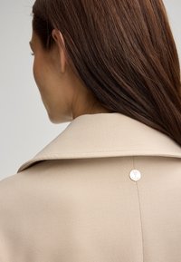 Beige op maat gemaakte jas met een brede kraag, voorzien van een zilveren cirkelvormig logo-accent en een gladde, gestructureerde stof.