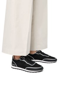 Schwarze Netz-Sneaker mit weißen Akzenten und Schnürsenkeln, mit abgerundetem Zehenbereich und strukturierter Sohle. Kombiniert mit cremefarbigen, weiten Hosen.
