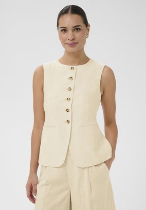Kvinde iført ærmeløs cremefarvet vest med knapper og matchende bukser, stående med den ene hånd i lommen mod en ensfarvet baggrund.