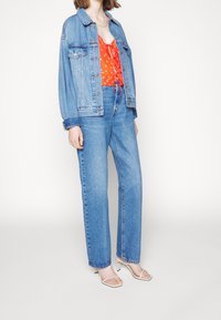 Femme portant une veste en denim bleu clair et un jean, un haut rouge à pois blancs, des sandales beige à talons hauts à brides, debout devant un fond blanc.