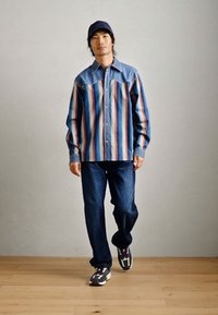 Ραβδωτό τζιν πουκάμισο με μπλε λεπτομέρειες, συνδυασμένο με σκούρα τζιν και αθλητικά παπούτσια. Το πουκάμισο διαθέτει κλασικό σχέδιο με κουμπιά.