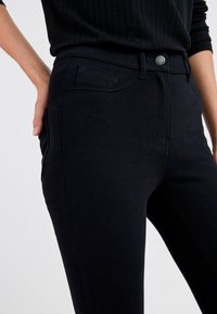 Jean en denim noir avec une coupe ajustée, doté d'une poche avant, de détails de coutures discrètes et d'une fermeture par bouton métallique à la taille.