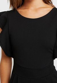 Svart klänning med korta, fladdriga ärmar, en bred rund halsringning och en slimmad överdel som övergår i en veckad kjol. Slät textur.