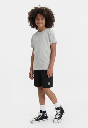 Copil cu păr creț purtând un tricou gri, pantaloni scurți negri cu un mic logo, șosete albe și adidași negri cu glezna înaltă, stând pe un fundal alb.