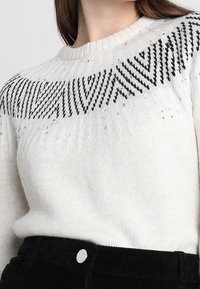 Vitt stickat tröja med ett svart geometriskt mönster längs yocken, rund halsringning och texturerad tyg. Kombinerat med svarta kordbyxor.