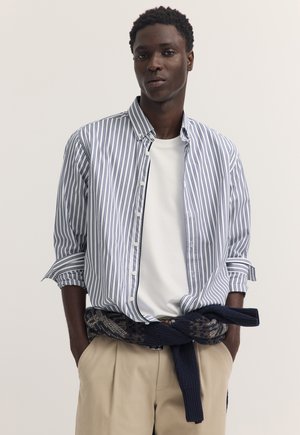 Junger Mann mit dunkler Haut und kurzen lockigen Haaren, der ein weißes T-Shirt, ein blau-weiß gestreiftes Hemd mit Knöpfen, beige Hosen und einen um die Taille gebundenen Pullover trägt.