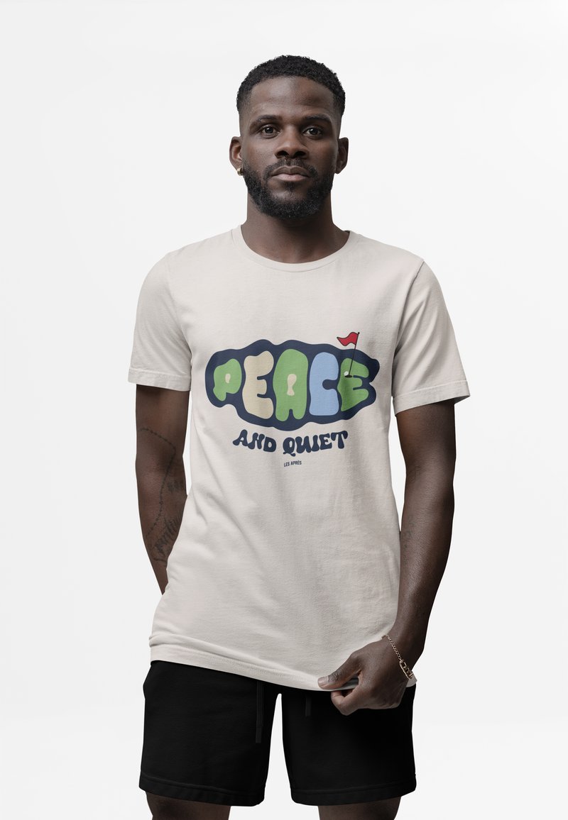 Mężczyzna w beżowej koszulce z kolorowym napisem "Pokój i cisza" oraz małą czerwoną flagą, w czarnych spodniach krótkich, stojący na białym tle.