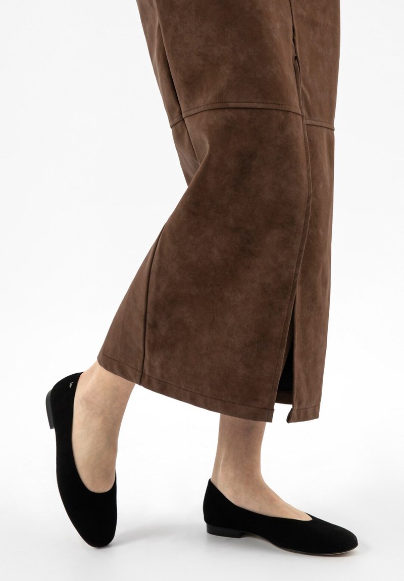 Gambe con gonna midi marrone con spacco laterale e scarpe nere chiuse con tacco basso su sfondo bianco.