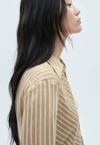 Camicia button-up color tan con righe verticali bianche, caratterizzata da un colletto a punta e maniche corte, mostrata sul profilo di un modello contro uno sfondo chiaro.