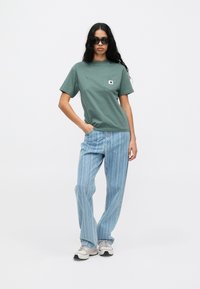 Roheline puuvillane t-särk rinna taskuga, koos helesiniste vertikaalsete triibuliste teksadega. Mudel kannab päikeseprille ja spordijalatu.