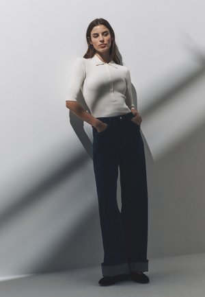 Mujer con cabello oscuro, vestida con una camiseta blanca de manga corta acanalada y vaqueros oscuros de pierna ancha, de pie con las manos en los bolsillos frente a una pared blanca.