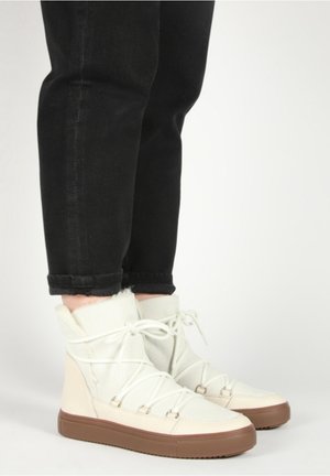 Osoba w beżowych, zimowych butach za kostkę z futrzanym wykończeniem i sznurowadłami krzyżującymi się, ubrana w czarne, podwinięte spodnie na białym tle.