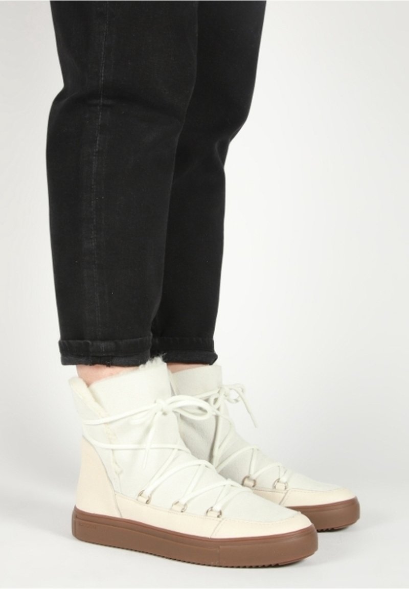 Personne portant des bottes d'hiver beige montantes avec une doublure en fourrure et des lacets croisés, associées à un pantalon noir retroussé sur un fond blanc.