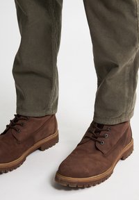 Botas de cuero marrón con ojales y cordones negros, superficie texturizada y suelas robustas, combinadas con pantalones de pana verde oliva.