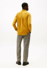 Uomo in piedi di lato, indossa una camicia gialla senape, pantaloni grigi a quadri e scarpe nere, su uno sfondo bianco uniforme.