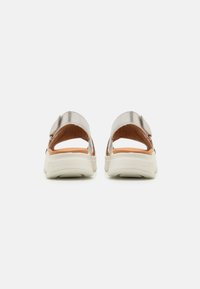 Achteraanzicht van lichtgekleurde sportsandalen met metallic banden, een gestructureerde bruine voetbed en een stevige witte zool.