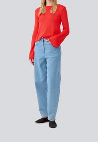 Pull en maille rouge à manches longues avec larges poignets, associé à un jean taille haute bleu clair à jambes droites. Des chaussures noires à enfiler complètent la tenue.