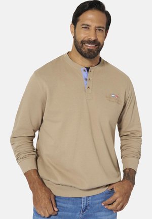 Man with beard wearing beige long-sleeve henley shirt with blue button placket and small embroidered flags, paired with blue jeans.