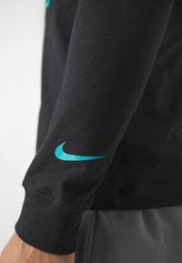Black long-sleeve shirt with a smooth texture, featuring a turquoise Nike logo on the left sleeve. Simple, fitted design.