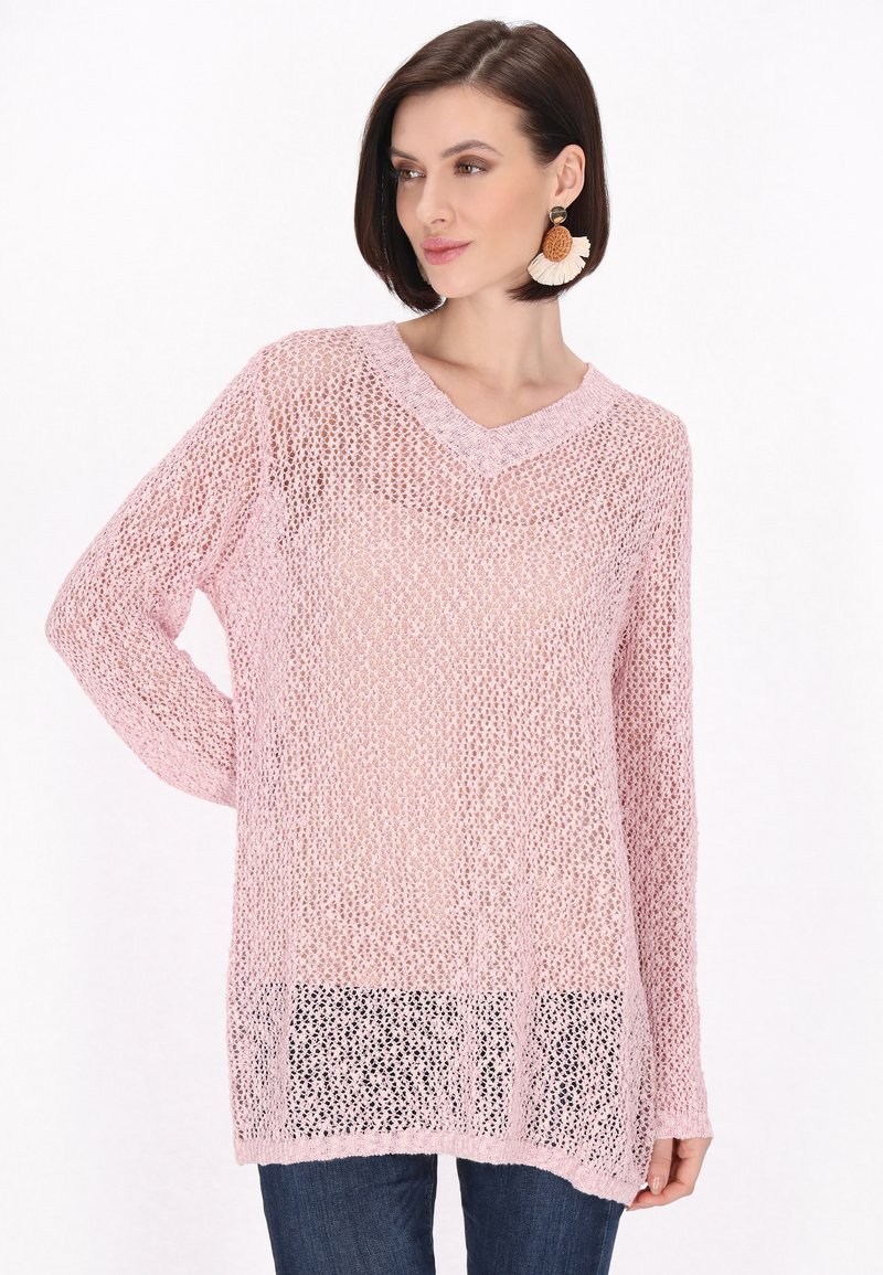 Hellrosa gestrickter Pullover mit V-Ausschnitt, langen Ärmeln, loser Struktur und leicht verlängertem Rücken, kombiniert mit dunklen Jeans.