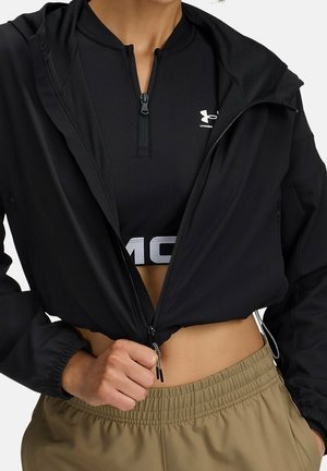 Person iført en sort lynlåstrøje over en sort Under Armour sportstop og beige bukser med elastik i taljen, trækker lynlåsen på jakken ned med hånden.