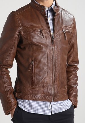 Homme portant une veste en cuir marron avec des poches zippées par-dessus une chemise à carreaux bleue et blanche, les mains partiellement dans les poches du pantalon.