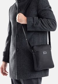 Borsa a tracolla nera realizzata in materiale resistente, con una superficie texturizzata e una cintura regolabile, mostrata su un cappotto grigio.