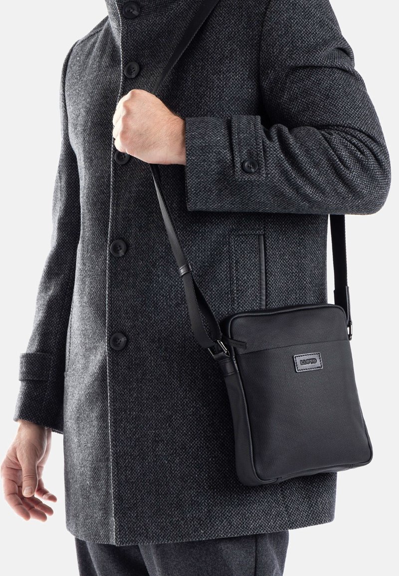 Borsa a tracolla nera realizzata in materiale resistente, con una superficie texturizzata e una cintura regolabile, mostrata su un cappotto grigio.