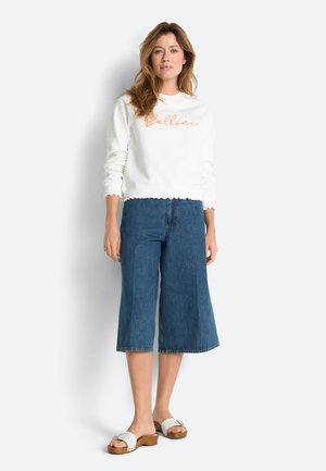 Frau steht und trägt ein weißes Langarmoberteil mit orangefarbenem Text, weit geschnittene, gekürzte blaue Jeans und weiße Sandalen mit Absatz.