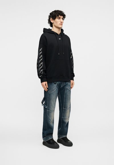 Jauns vīrietis, tērpies melnā kapučjakā ar apdrukātām piedurknēm, zilās džinsa darba biksēs un melnās sporta kurpēs, stāv pret vienkāršu gaišu fonu.