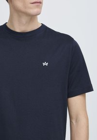 Marineblauwe katoenen t-shirt met een ronde halslijn en korte mouwen. Bevat een klein wit geborduurd logo op de borst. Glad textuur.