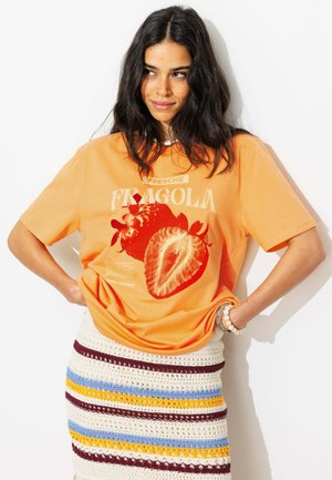 Femme aux longs cheveux foncés porte un t-shirt orange avec un imprimé fraise rouge et un texte, assorti à une jupe blanche à rayures bleues, jaunes et bordeaux.