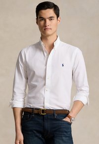 Witte lange mouwen overhemd met knoopsluiting en een kraag, voorzien van een klein blauw logo, gecombineerd met donkere jeans en een bruine riem. Strak geweven stof.