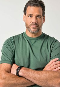 Green short-sleeve t-shirt with a round neckline, textured fabric, and decorative stitching. Black leather bracelet on the wrist.