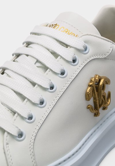 Λευκό αθλητικό παπούτσι με χοντρές λευκές κορδόνια, λογότυπο με ανάγλυφο χρυσό στη πλευρά και χρυσό κείμενο με το όνομα της μάρκας στη γλώσσα.